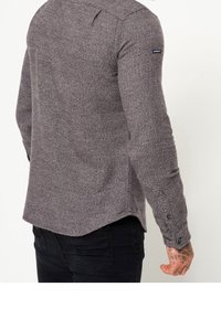 Camisa de manga larga gris texturizada con dobladillo curvado, puños abotonados y un pequeño parche de logo en la parte superior del brazo. La tela muestra un patrón sutil.