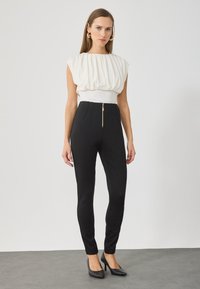 Λευκή, φούτερ μπλούζα με ψηλό λαιμό, σε συνδυασμό με ψηλόμεσα μαύρα παντελόνια με μπροστινό φερμουάρ και κομψή γραμμή. Μαύρες γόβες.