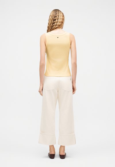 Γυναίκα με μακριά κυματιστά μαλλιά, ντυμένη με κίτρινο αμάνικο τοπ, λευκό παντελόνι φαρδιάς γραμμής και σκούρα καφέ παπούτσια, στέκεται με την πλάτη γυρισμένη.