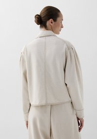 Beige cotton blend jacket with detailed stitching, relaxed fit, and shirt collar, shown from the back with slightly puffed sleeves.