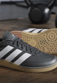 Zwart-witte Adidas sneakers met rubbere zolen op een houten vloer, fitnessgewichten wazig op de achtergrond.