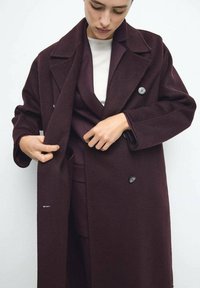 Bordeauxroter Wollmantel mit doppelreihigem Design, großen Revers und zwei Knöpfen. Das Modell richtet den Mantel zurecht und betont dessen Textur und Passform.