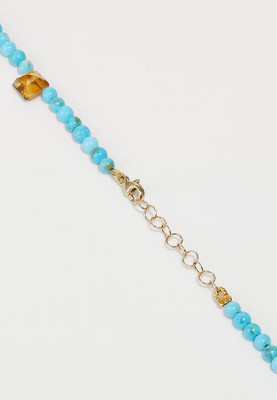 Μια βραχιόλι με κοκκάλια σε τιρκουάζ χρώμα που διαθέτει χρυσές λεπτομέρειες και κούμπομα με καβίλιο. Περιλαμβάνει μια ξεχωριστή χάντρα σε πορτοκαλί χρώμα ανάμεσα στις τιρκουάζ.