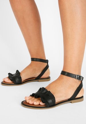 Des pieds portant des sandales à plat noires avec des brides de cheville et des brides avant en cuir froncé sur un fond clair.