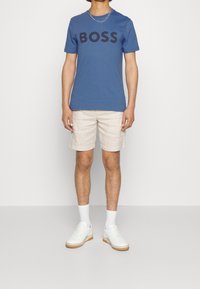 Blå bomullströja med svart "BOSS"-logotyp, kombinerad med ljusbeiga shorts. Vita sneakers med gummisålar och vita ankelsockor fulländar looken.