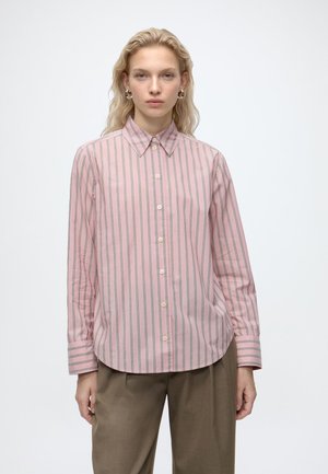 Vrouw met licht haar draagt een roze blouse met lange mouwen met verticale strepen en taupekleurige broek, staand tegen een effen witte achtergrond.