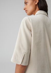 Camicia in corduroy beige chiaro con colletto, maniche corte e finitura testurizzata. Il design presenta una sottile rigatura verticale.