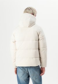 Chaqueta de plumas en beige claro con capucha, que presenta un diseño acolchado y un corte recto. Combinada con vaqueros azules, mostrando una textura neutra.