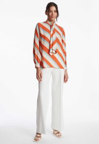 Mehrfarbige Bluse mit lebhaften orangefarbenen, blauen und cremefarbenen diagonalen Streifen; verfügt über einen Krawattenkragen und lange Ärmel, kombiniert mit weißen geraden Hosen.