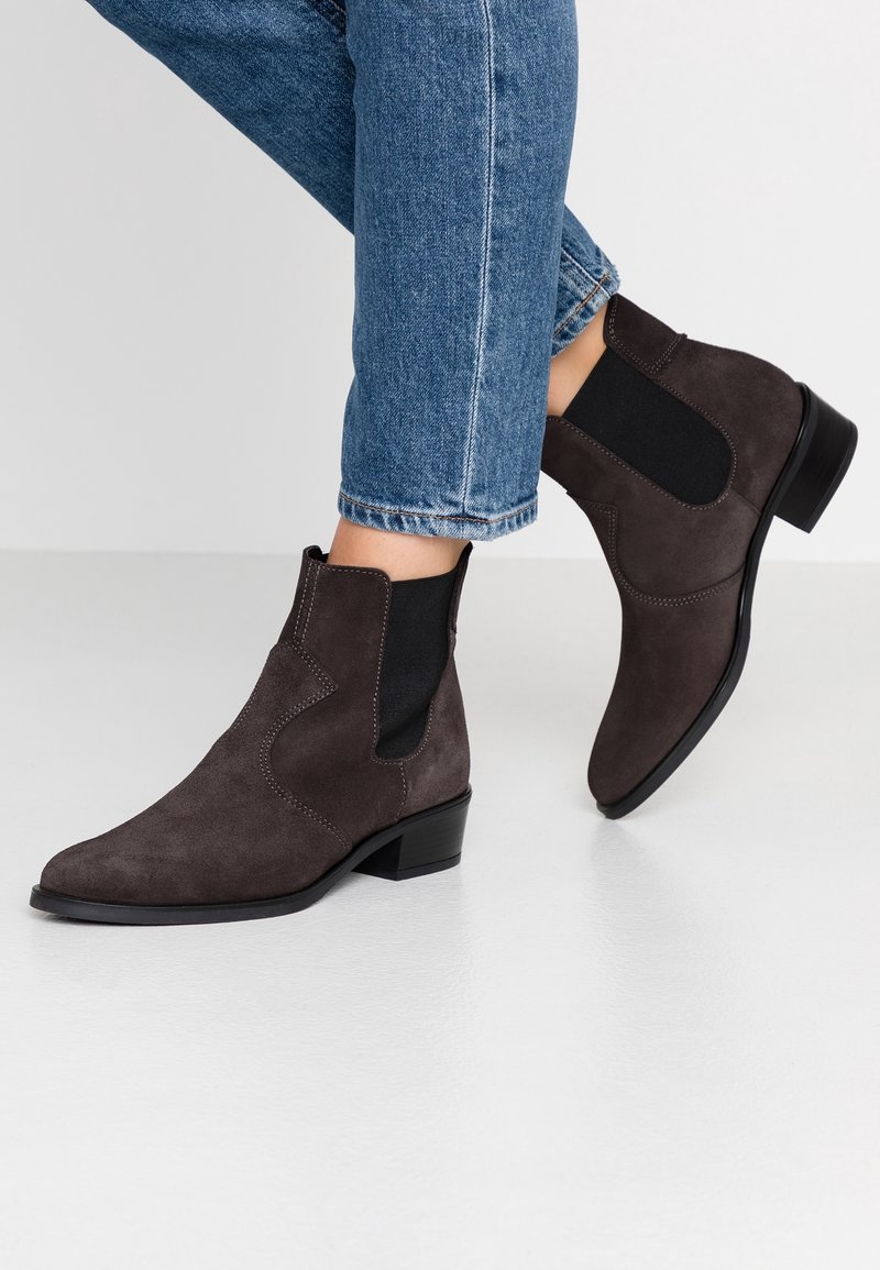 Bruine suède enkellaarsjes met elastische zijpanelen, een puntige neus, lage blokhak en contrasterende stikseldetails. Gecombineerd met een blauwe spijkerbroek.