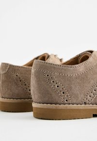 Chaussures en daim marron présentant des perforations décoratives, un bout rond et une semelle en caoutchouc avec un détail de couture visible sur le bord.