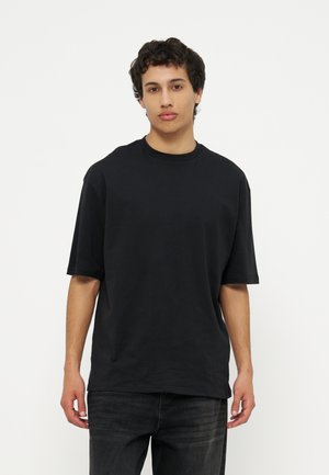 Young man with curly dark hair wears a loose black T-shirt and black jeans, standing against a plain white background.