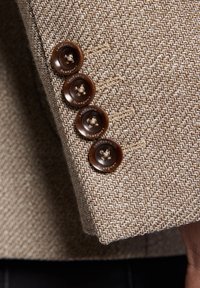 Brązowy materiał marynarki o teksturze w jodełkę z czterema ciemnobrązowymi guzikami z wygrawerowaną marką na mankiecie.