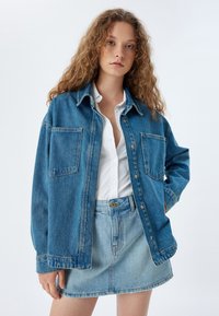 Jeansjacka med två framfickor, ljusblå nyans, avslappnad passform; tillsammans med en matchande jeanskjol. Vit skjorta med knappar under.