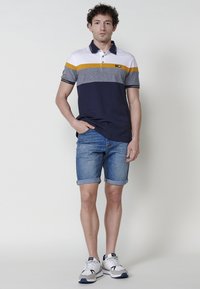 Polo majica sa plavim i sivim prugama, žutim akcentom, kratkim rukavima i kragnom. Nosi se uz poredane traper kratke hlače i sportske tenisice.