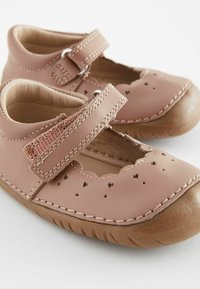 Różowe skórzane buty z perforowanymi wzorami w kształcie serca, regulowanym paskiem, falbaniastymi krawędziami i brązową gumową podeszwą. Miękka, zaokrąglona konstrukcja palców.