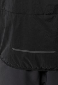 Giacca a vento nera con una texture liscia e leggera, orlo elasticizzato e un dettaglio riflettente vicino al fondo.