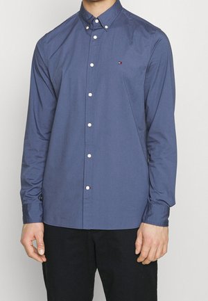 Uomo che indossa una camicia elegante blu scuro a maniche lunghe con bottoni bianchi e un piccolo logo sul petto, abbinata a pantaloni neri.