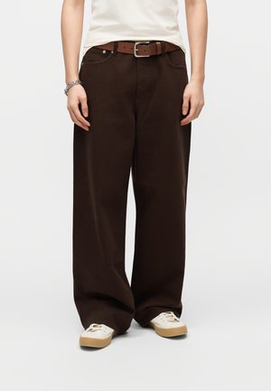 Person iført løstsiddende mørkebrune bukser, hvide sneakers med gummisåler, et brunt bælte og et sølvarmbånd på venstre håndled.