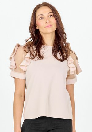 Mujer con cabello largo y castaño, vestida con una blusa beige con mangas de volantes en capas y pantalones negros, posando frente a un fondo blanco.