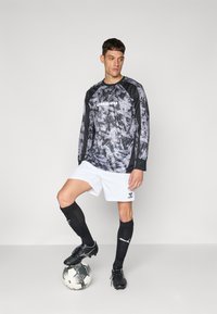 Langärmliges graues Camouflage-Trikot mit schwarzen Akzenten, weißen Shorts, schwarzen Socken und schwarzen Fußballschuhen auf einem Fußball.