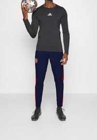 Musta pitkähihainen urheilupaita, jossa valkoinen logo, yhdistettynä tummansinisiin housuihin, joissa on punaiset raidat ja Arsenal-logo. Mustat jalkapallokengät.