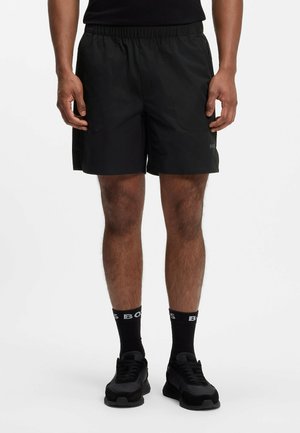 Mand iført sorte atletikshorts, sorte sokker med hvid tekst og sorte sneakers, stående mod en ensfarvet baggrund.