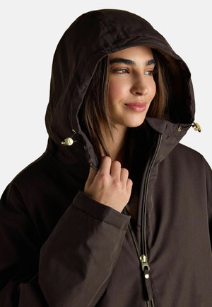 Frau in einer dunkelbraunen Kapuzenjacke, die die Kapuze nahe ihrem Gesicht hält, nach rechts schaut und leicht lächelt.