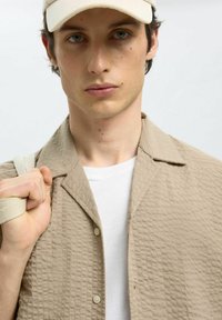 Lichtbeige geborduurd shirt met een korte kraag, knoopsluiting aan de voorkant, over een wit T-shirt; persoon houdt een riem met een crèmekleurige accent.