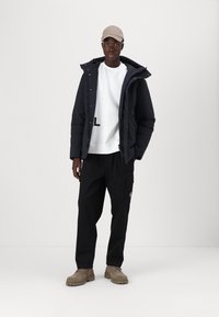Sort pufferjakke, hvid sweatshirt med tryk, sorte cargo-bukser og beige kasket. Brune sko med strukturerede såler fuldender outfittet.