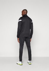 Jachetă sportivă neagră cu accente albe pe umeri și pantaloni de trening asortați. Are guler cu fermoar, material elastic și un design într-o croială ajustată.