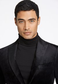 Zwart fluwelen tuxedo jasje met een soepele textuur, voorzien van een sjaalkraag en een borstzak, gecombineerd met een strakke zwarte turtleneck.