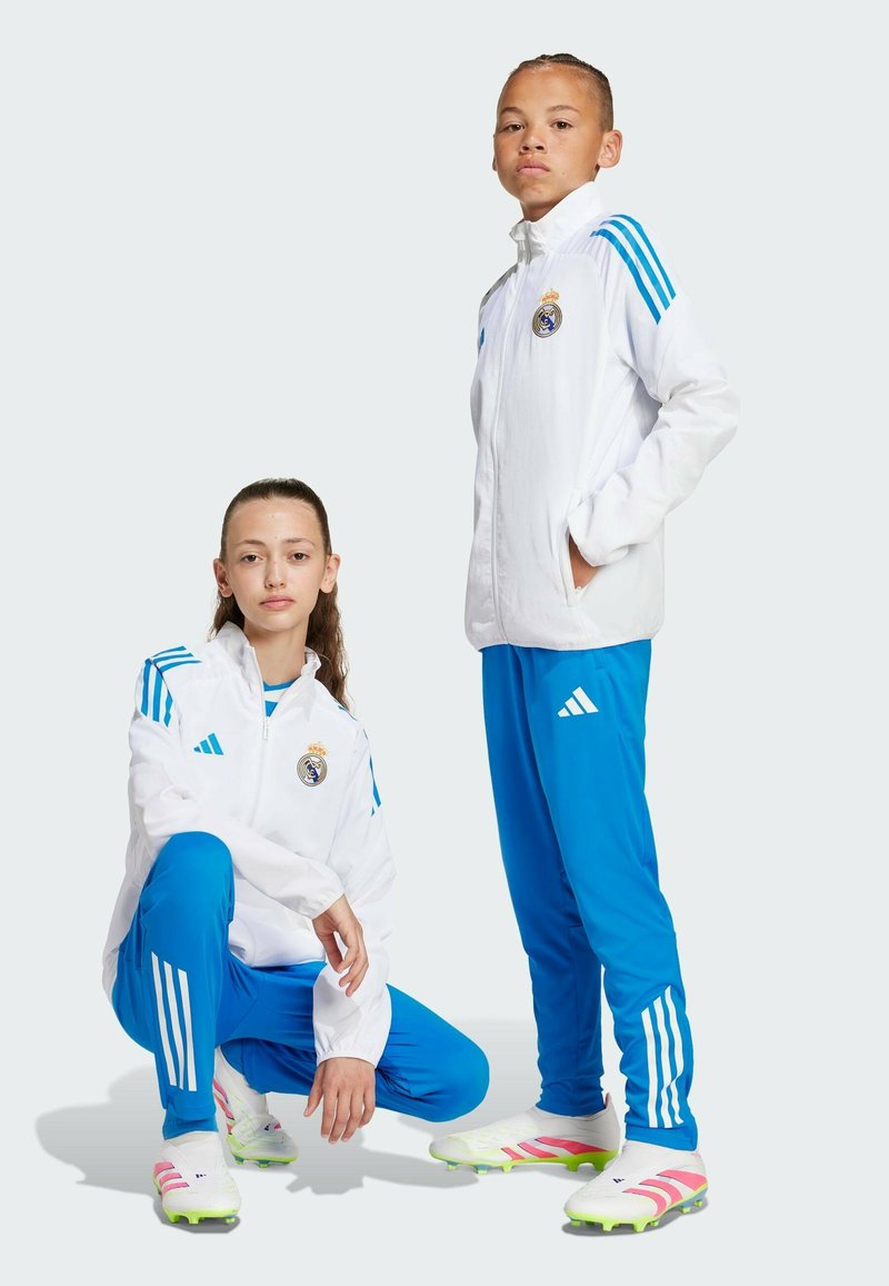 Weiße Sportjacken mit blauen Streifen und dem Real Madrid-Logo, kombiniert mit blauen Hosen und weißen Fußballschuhen mit rosa Akzenten, vor einem hellen Hintergrund.