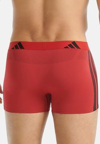 Červené sportovní kraťasy se přiléhavým střihem. Mají černé pruhy a logo na pase. Materiál je hladký s mírným leskem.