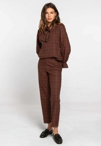Conjunto de cuadros marrones con una blusa que presenta un lazo y mangas sueltas, combinado con pantalones ajustados. El material parece suave, con una textura lisa.
