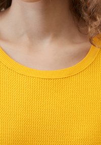 Femme aux cheveux bouclés portant un pull jaune texturé, visible des épaules au cou sans visage visible.