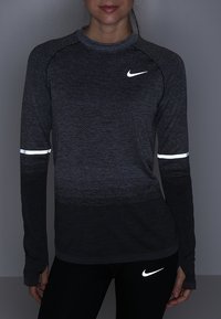Dlouhá sportovní tričko Nike v tmavě šedé barvě s texturovaným vzorem, reflexními detaily na rukávech a přiléhavým střihem.