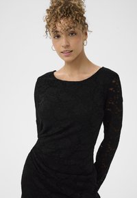 Crna čipkasta haljina s dugim rukavima, okruglim izrezom, prilagođenim tijelom i naboranom detaljnom obradom u struku. Glatka tekstura, vidljiv cvjetni uzorak.