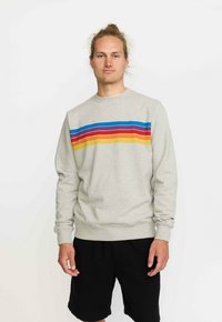 Jonge man met lichtbruin haar in een bun, gekleed in een lichtgrijze sweatshirt met meerkleurige horizontale strepen en zwarte shorts, staande tegen een witte achtergrond.