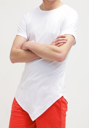 Mann trägt ein schlichtes weißes T-Shirt mit kurzen Ärmeln und asymmetrischem, spitz zulaufendem Saum sowie leuchtend roten Shorts, die Arme verschränkt.