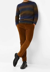 Homme portant un pull dégradé bleu et marron, un pantalon en velours côtelé couleur rouille, des chaussures habillées noires, debout les mains dans les poches.