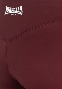 Shorts de sport bordeaux en tissu lisse et extensible, avec des coutures élastiques et un logo Lonsdale blanc bien visible.