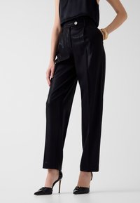 Frau trägt schwarze, hoch taillierte, satinierte, zulaufende Hosen, ein ärmelloses schwarzes Oberteil, schwarze spitze High Heels und ein goldenes Armband an einem Handgelenk.