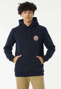 Marineblauwe hoodie met een trekkoord, een voorzak en een cirkelvormig roze logo op de borst. Zachte stof met ribgebreide manchetten en onderrand.