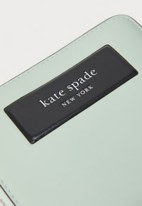 Mintgrön läderplånbok med en svart läderetikett med präglad text "kate spade NEW YORK." Slät textur med synliga sömmar.