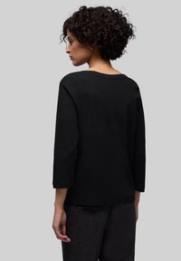 Kobieta o kręconych włosach, ubrana w czarną, długorękawową bluzkę i ciemne spodnie, zwrócona bokiem na jednolitym szarym tle.