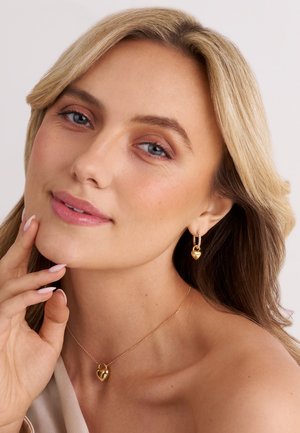 Guld hjärtformade örhängen och matchande halsband på en person med mjuka vågor i håret. Slät hud och naturlig makeup, med ljusa naglar.