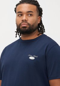 Tamsiai mėlynas medvilninis marškinėlis su apvalia apykakle ir trumpomis rankovėmis, ant krūtinės baltai siuvinėtas logotipas, lygios tekstūros ir laisvo modelio.