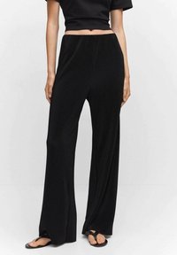 Femme portant un pantalon large côtelé noir, un top court noir et des sandales noires, debout devant un fond gris uni.