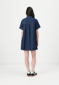 Denim jurk in donkerblauw, korte mouwen, met knoopsluiting aan de kraag en een losse pasvorm. Opvallende stiksels en een gebogen zoomlijn.